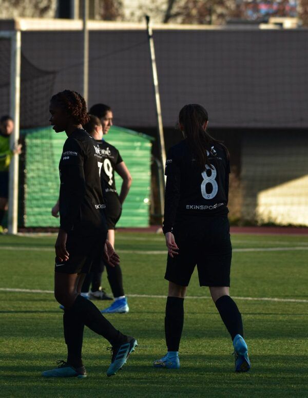 Un groupe de joueuses de football portant des uniformes noirs avec des numéros blancs se tiennent sur un terrain de football, discutant peut-être du match. L'arrière-plan montre un gardien de but près du but, une clôture et quelques bâtiments. Comment nous personnalisons les maillots pour notre équipe de foot féminine ?