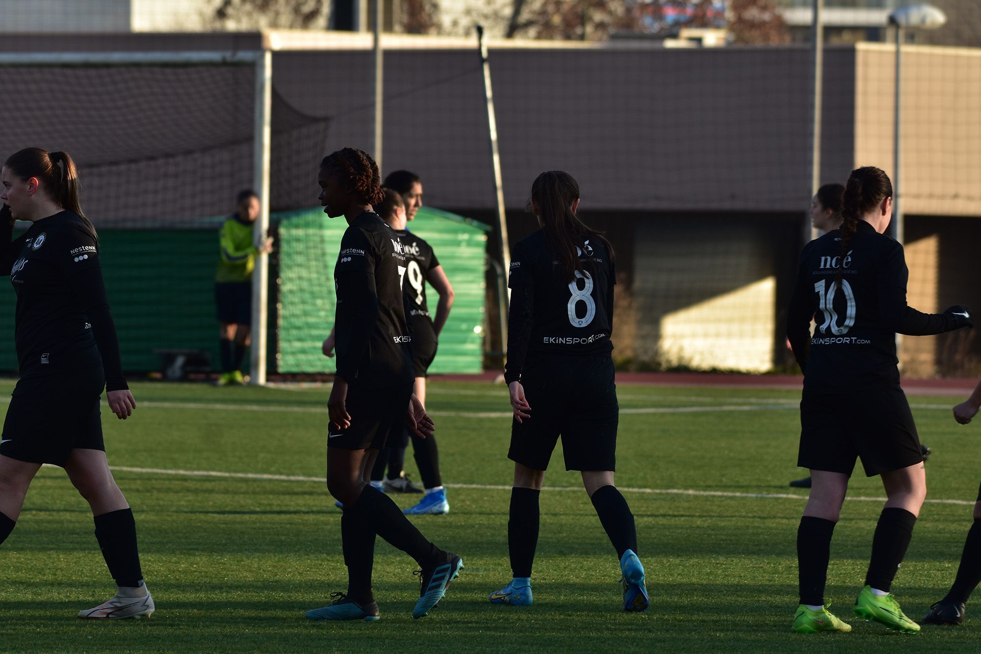 Un groupe de joueuses de football portant des uniformes noirs avec des numéros blancs se tiennent sur un terrain de football, discutant peut-être du match. L'arrière-plan montre un gardien de but près du but, une clôture et quelques bâtiments. Comment nous personnalisons les maillots pour notre équipe de foot féminine ?