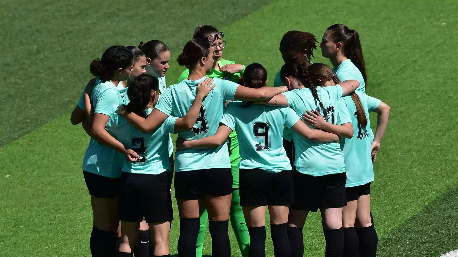 Une équipe de football féminin en maillot bleu clair et short noir se rassemble sur un terrain gazonné, faisant preuve d'unité et de travail d'équipe. La gardienne, vêtue d'un uniforme vert, est au centre du groupe. Le soleil brille de mille feux, projetant des ombres distinctes sur l'herbe.